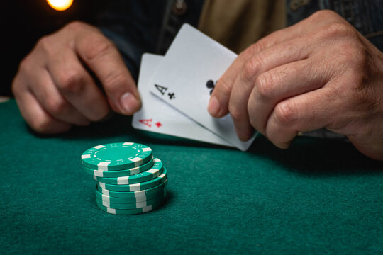 A man sits at a green table with poker chips stacked in front of him. He holds two playing cards with aces on them. The action takes place in a night casino