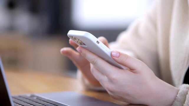 Person holds smartphone while laptop sits open nearby. Fingers tap screen. Hands wear soft sweater. Background blurred. Indoor workspace setting