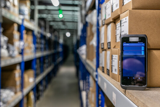 Logistics e-commerce warehouse interior featuring multi-tier shelving and pallet racking, with a handheld barcode scanner or rugged mobile computer placed on a storage shelf for inventory tracking