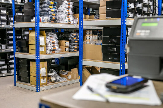Logistics e-commerce warehouse interior featuring multi-tier shelving and pallet racking, with a handheld barcode scanner or rugged mobile computer placed on a packing bench beside a label printer