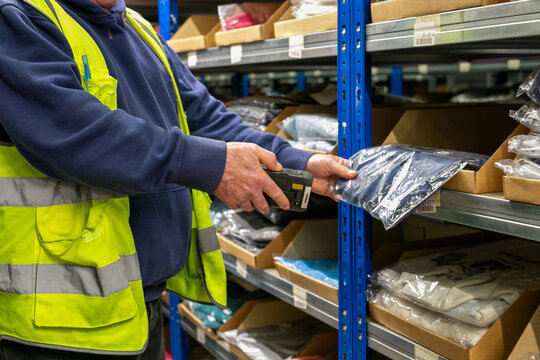Logistics warehouse operative wearing a high-visibility yellow vest and using handheld barcode scanner to scan labels on an item while performing inventory picking tasks in an inventory storage aisle 