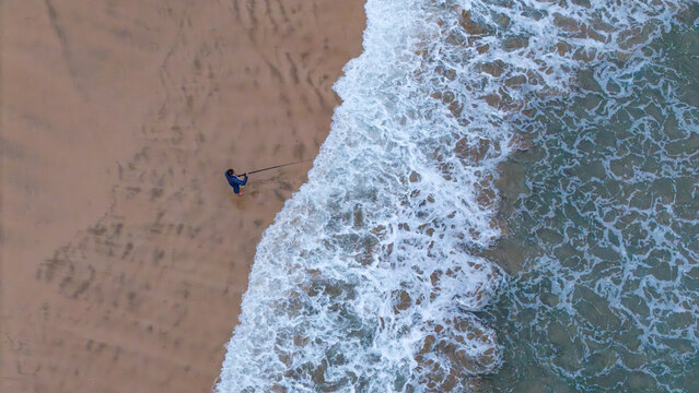 Aerial view of a lone fisherman standing on the sandy shore of Hartenbos beach with white waves crashing against the coast Mossel Bay, Western Cape, South Africa.