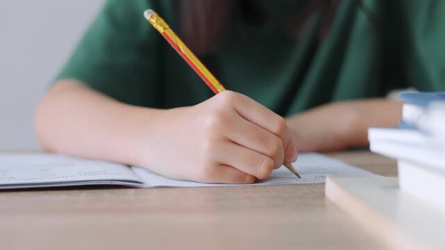 Close up selective focus of a student's hand holding a pencil, writing in a notebook. Bright daylight for the workspace, representing academic concentration,homework, and childhood education concepts.