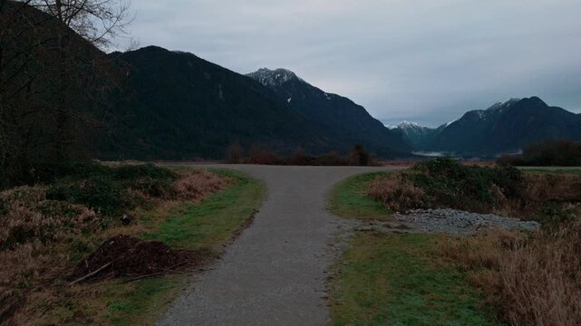 Drone glides along gravel trail over dark marshwaters toward towering snow mountains heavy overcast sky reflecting peaks in calm wetland waters sedge grasses muddy banks winter atmosphere approaching