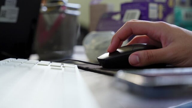 A close-up shot of a person's hand using a black computer mouse on a busy office desk. The footage captures digital workflow and professional productivity in a modern indoor workspace.