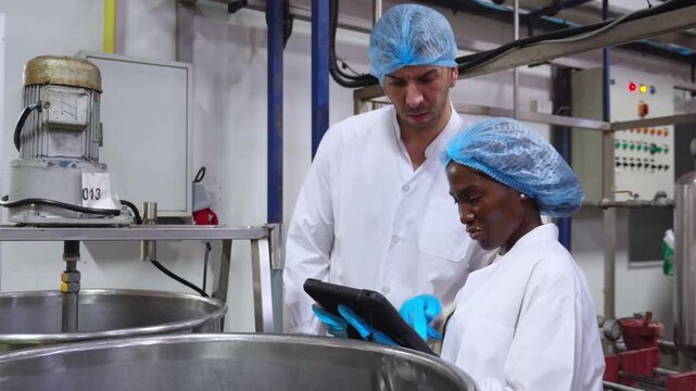 Diverse factory team workers inspecting large metal tank, Caucasian man African American woman using digital tablet checking manufacturing, food processing plant production line machinery.