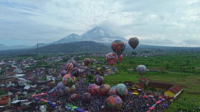 Large hot air balloon festival with colorful balloons and spectators. Wide drone shot over countryside landscape. Wonosobo balloon festival, Indonesia.