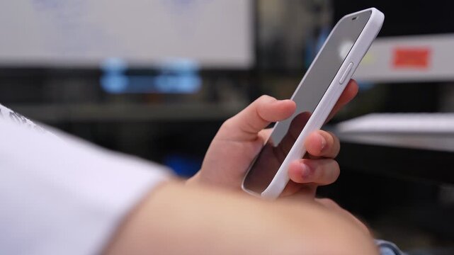 A person holds a white smartphone in a modern indoor workspace. The blurred background shows an office desk with a computer monitor and keyboard during daylight.