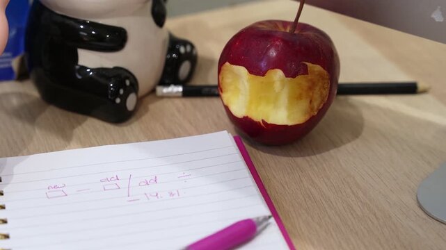 A bitten red apple sits next to a spiral notebook containing a handwritten math formula on a wooden desk. A pink pen and panda figurine create a relatable student or home office workspace.