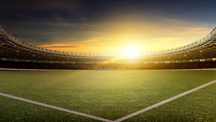 Fototapeta premium A wide-angle view of a professional football stadium at sunset. The empty arena features a lush green field under a dramatic golden hour sky with a bright lens flare