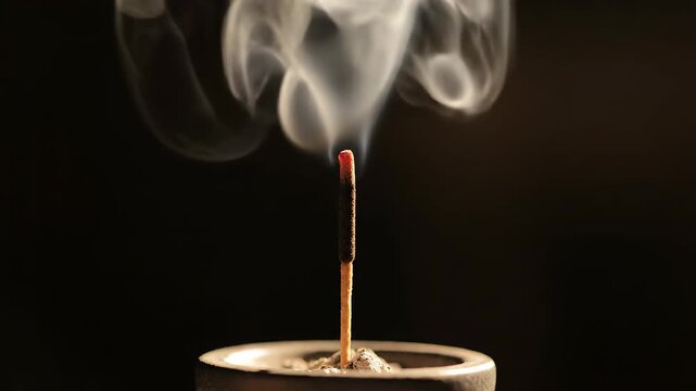 a single lit incense stick. Thin white smoke rises slowly and curls from the glowing tip against a very dark background. Minimal warm lighting. No hands.