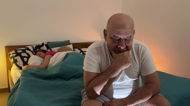 Hombre preocupado sentado en la cama mientras su pareja duerme