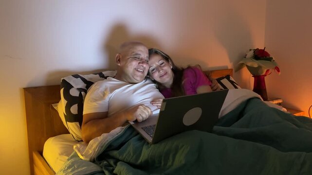 Pareja abrazada en la cama riendo mientras mira el port&aacute;til