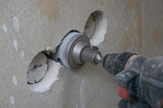 Worker drilling a large circular hole in a rough concrete wall using an electric hammer drill with a core bit, creating openings for electrical boxes during renovation