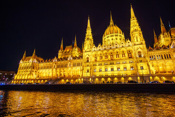 Fototapeta premium Illuminated Hungarian Parliament Building at Night on the Danube River in Budapest, Hungary, Featuring Gothic Revival Architecture and Golden Lights Reflecting in Water