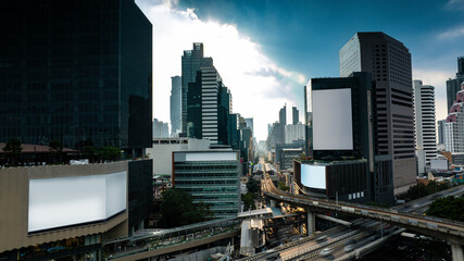 Fototapeta premium Wide aerial view of Bangkok’s modern business district featuring elevated BTS skytrain tracks, skyscrapers, urban traffic, and large blank billboard mockup spaces under dramatic sunset light. Ideal fo