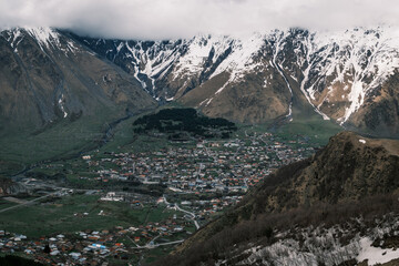 Obraz premium Panoramic view of Stepantsminda village nestled in the Caucasus Mountains near the Russian border, Georgia. Alpine settlement under snowy peaks and low clouds in the Kazbegi region