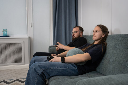 Young couple sitting on a living room couch, completely focused on playing video games with controllers, enjoying indoor entertainment and leisure time together