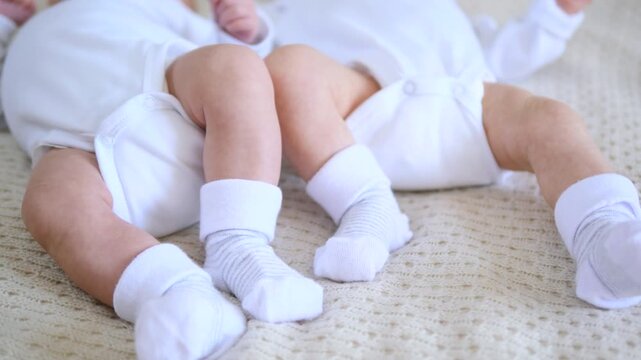 Adorable newborn twin babies in white bodysuits and socks kicking their tiny feet while lying together on a soft, knitted blanket, representing innocence and new life in early childhood
