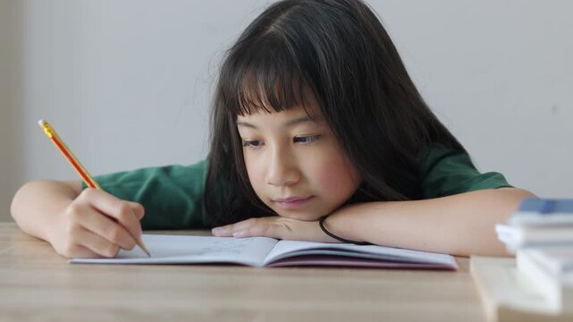 Close up selective focus of demotivated Asian kid girl resting her head on a desk in daylight. She looks overburdened by too much homework, illustrating academic pressure and childhood study burnout.