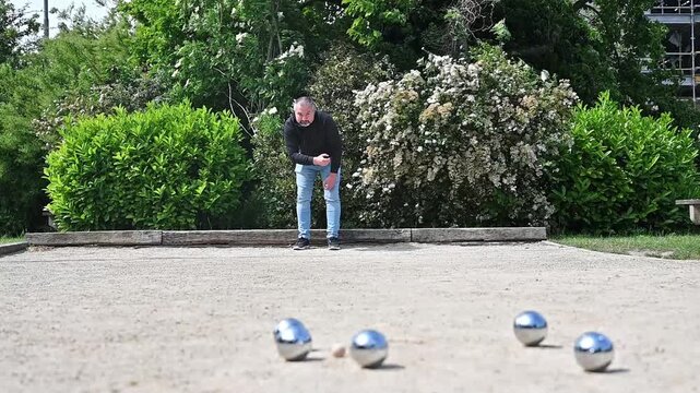 Slow motion of man tossing steel petanque ball on park court with trees behind, graceful throwing arc and outdoor boules game movement