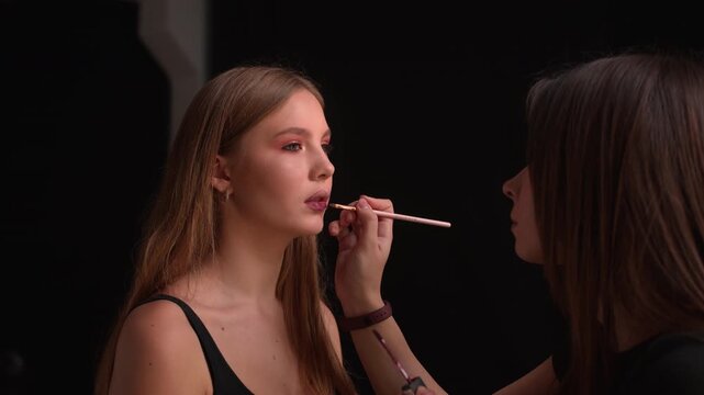 Professional makeup artist applying dark lipstick on a beautiful model's lips in a beauty salon, creating a professional and fashionable look against a black background