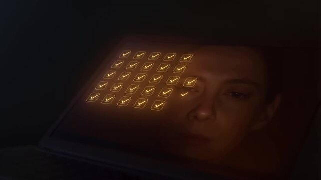 A woman stares intently as a grid of golden checkmarks fills her screen, her reflection visible in the dark monitor.