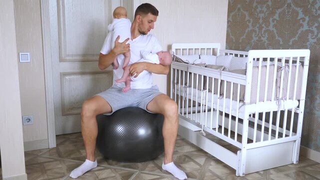 Loving father sitting on a fitball while holding his two newborn twin babies, gently rocking and lulling them to sleep in a cozy nursery room next to a white crib with tender care