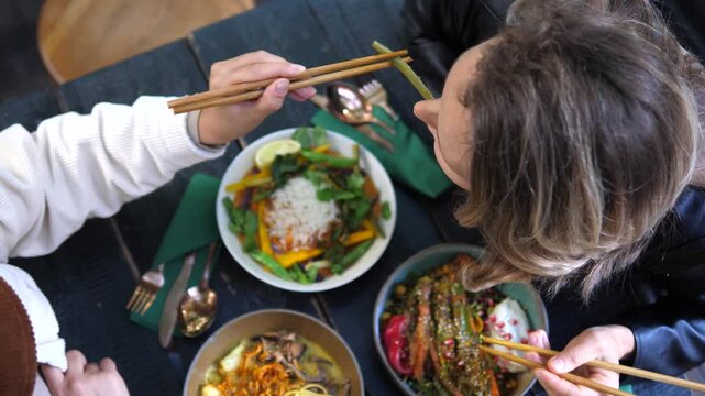 Top down view of two women enjoying a healthy and colorful vegan lunch at a dark wooden table, using chopsticks to eat various plant based dishes from beautiful ceramic bowls