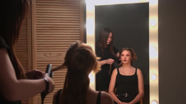 Professional hairstylist creating an evening hairstyle for a beautiful young woman using a curling iron in a beauty salon with a large mirror and warm lights