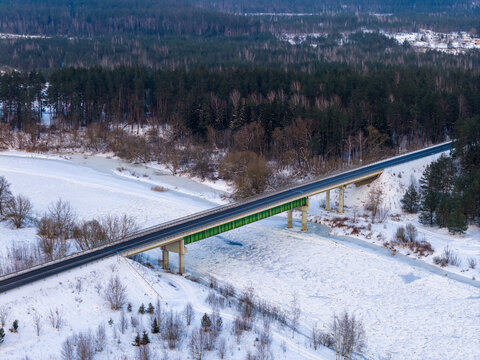 Aerial view of a bridge crossing a frozen river surrounded by snow-covered banks and a dense evergreen forest Lithuania.