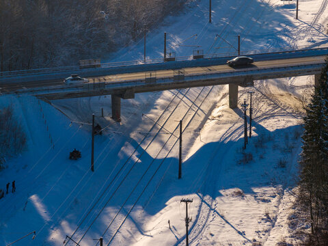 Aerial view of a road bridge crossing over snow-covered railway tracks with people and quad bikes in a wintry forest with long shadows in Lithuania.