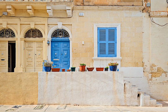 Sunny facade of historic Malta limestone building with bright blue wooden door, matching shutters, ornate ironwork arch, terracotta and blue flower pots on white wall