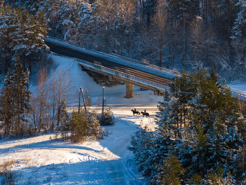 Aerial view of a bridge over a snowy path with two horse riders surrounded by frost-covered pine trees in Lithuania.