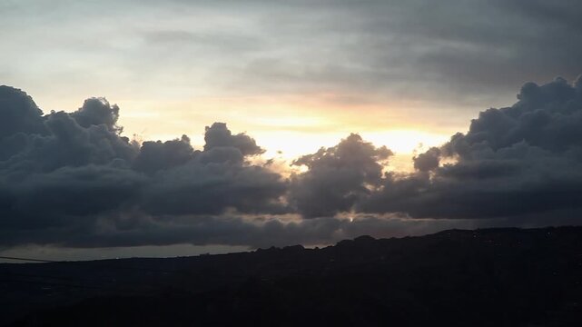 Pan of the sky at sunset, full of intertwined clouds, in the middle the sun hides forming a golden space in a grayish-blue sky.
