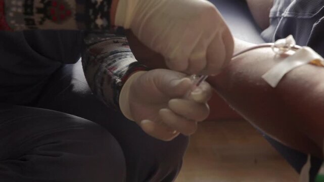 A nurse draws blood into glass test tubes from a man. Wearing rubber gloves, she transfers the blood from a plastic tube into the test tubes with great expertise.