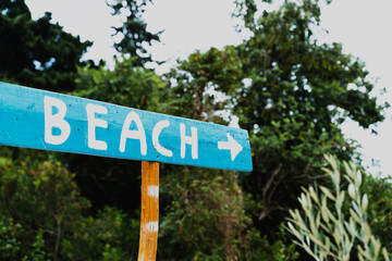 Fototapeta premium A blue wooden sign with white letters and an arrow points the way to the beach through a dark coastal forest. Summer vacation direction and secluded island travel concept.