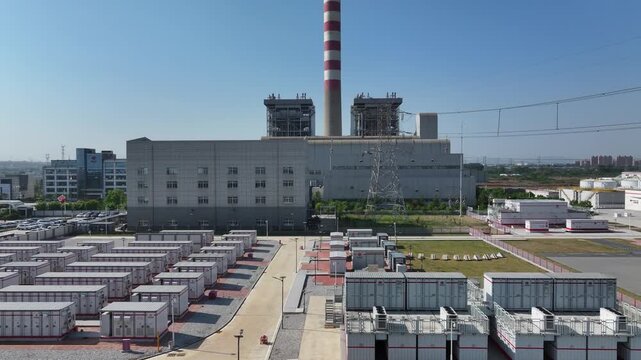 Thermal power plant facility with tall chimney, auxiliary buildings, and rows of electrical equipment under clear blue sky