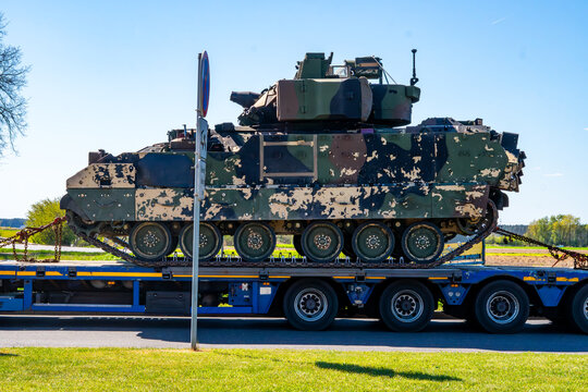 Side profile of an American Bradley infantry fighting vehicle in desert camouflage, transported on a multi-axle lowboy trailer, showing caterpillar tracks and armor details against trees and blue sky.