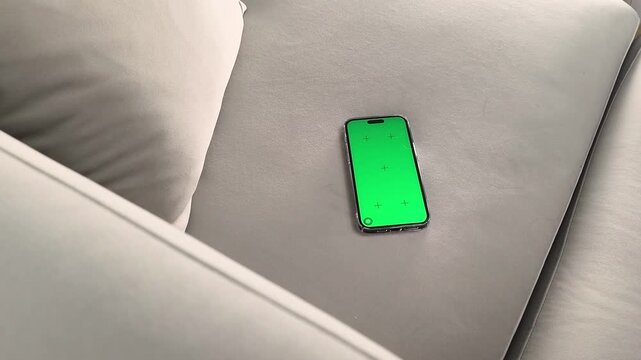 Smartphone with green screen lies on sofa and finger taps lower part of display. Hand presses on chroma interface at home. Concept mobile control, app interaction, digital usage, touch input