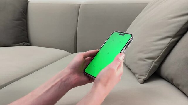 Hands hold smartphone with green screen and scroll on display while sitting on sofa. Fingers interact with chroma interface using touch gesture. Concept app mockup, mobile navigation,