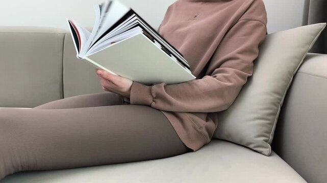 Woman leans on pillow browsing book pages while sitting on sofa. Female scans content quickly during relaxed home moment. Leisure reading and rest concept in apartment living room..