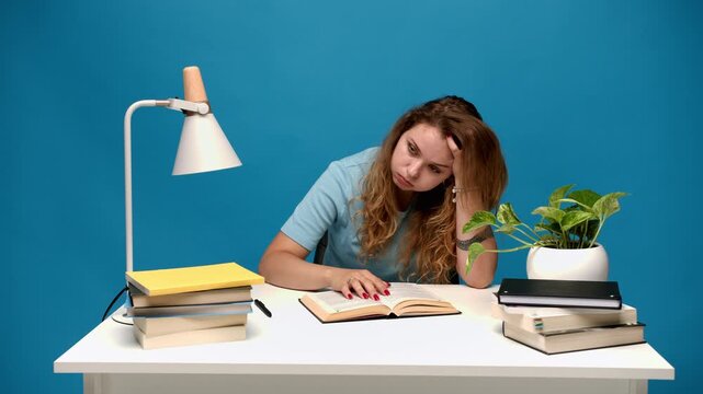 Tired female student feeling bored while studying hard at her desk for a difficult college exam surrounded by many books