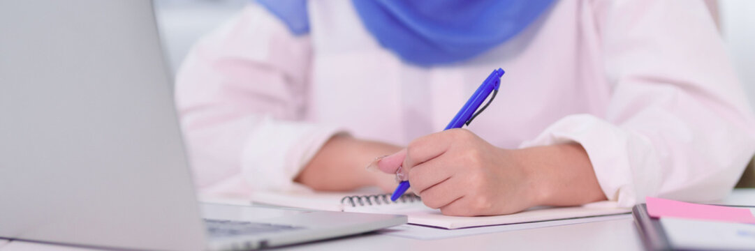 Muslim student women writing notes in notebook studying at desk with laptop computer for education learning business work at home office wearing hijab headscarf