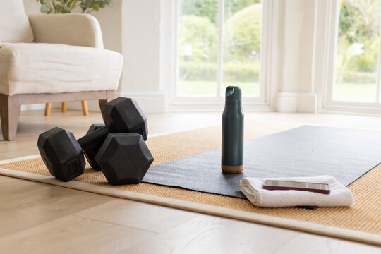 Yoga mat, dumbbells, teal bottle and towel holding phone are sitting by windows, sunlight streaming