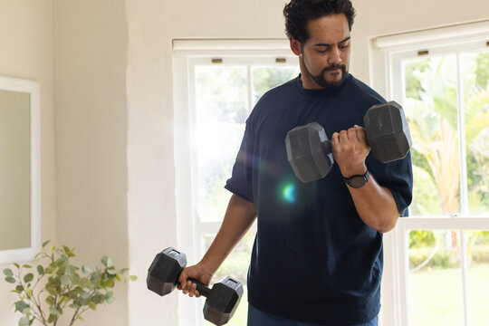 African American mid-adult man curling hex dumbbell near windows at home, wearing watch and gymwear
