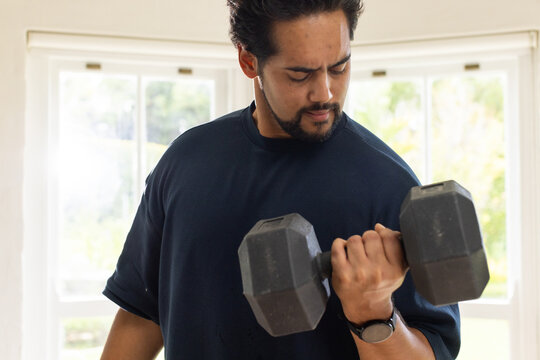 Mid-adult African man lifting hex dumbbell at home wearing navy tee and watch near white windows