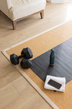Dumbbells and mat are resting on woven rug in living room corner, with towel, bottle, smartphone