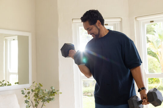 Middle-aged African American man lifting hex dumbbells while training in living area near windows