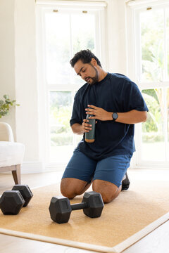 Midlife African American man kneeling in living room in workout gear twisting shaker near weights